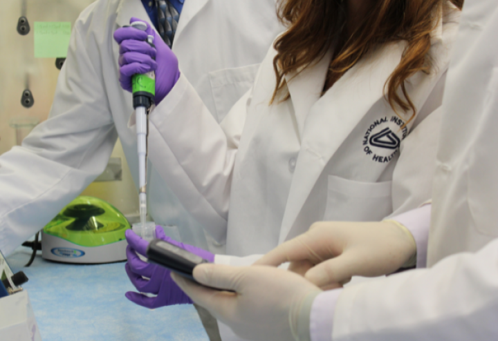 Two people in lab coats stand around a woman in a lab coat holding up a syringe. Source: National Institute of Health. 