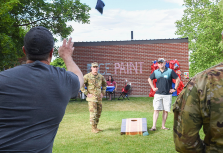 A man tosses a sack for a game of cornhole with a man in a miltary uniform on the other side and a Face Paint sign behind them